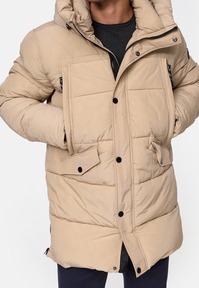 Veste d'hiver beige matelassée avec capuche, boutons noirs, poches à rabat, portée sur une chemise foncée, les mains glissées dans les poches latérales.