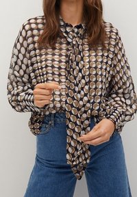 Blusa marrón con estampado de diseños circulares en tonos variados, con botones, de manga larga y un lazo en el cuello. Combinada con jeans de mezclilla.