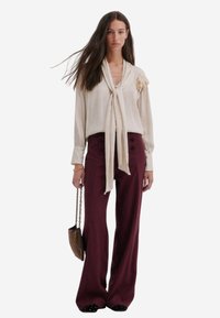 Beige blouse met lange mouwen en een gestrikte hals, afgewerkt met een bloemaccent, gecombineerd met hoog geïndekte bordeauxrode wijde broek met knopen. 