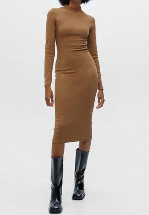 Femme portant une robe midi ajustée à manches longues de couleur camel associée à des bottes en cuir noires montantes jusqu’au genou sur un fond uni.