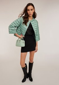 Jachetă cu croi scurt, cu model matlasat, de culoare verde mentă, cu nasturi tip snap, top negru transparent, fustă mini neagră, cizme negre până la genunchi și geantă de mână verde mentă.