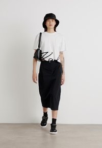 Hvit kortermet t-skjorte med svart tekst, svart midikjole, svart bøttehatt og svarte joggesko. Modell med en liten svart veske.