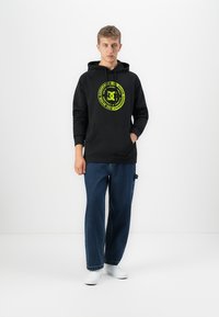 Young man standing wearing black hoodie with yellow DC logo, loose blue jeans, and white sneakers against white background.