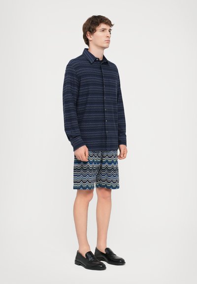 Jūrų tematikos ilgomis rankovėmis marškiniai, daugiaspalvės zigzago rašto šortai ir juodi slip-on tipo batai, modelis stovi prieš paprastą baltą foną.
