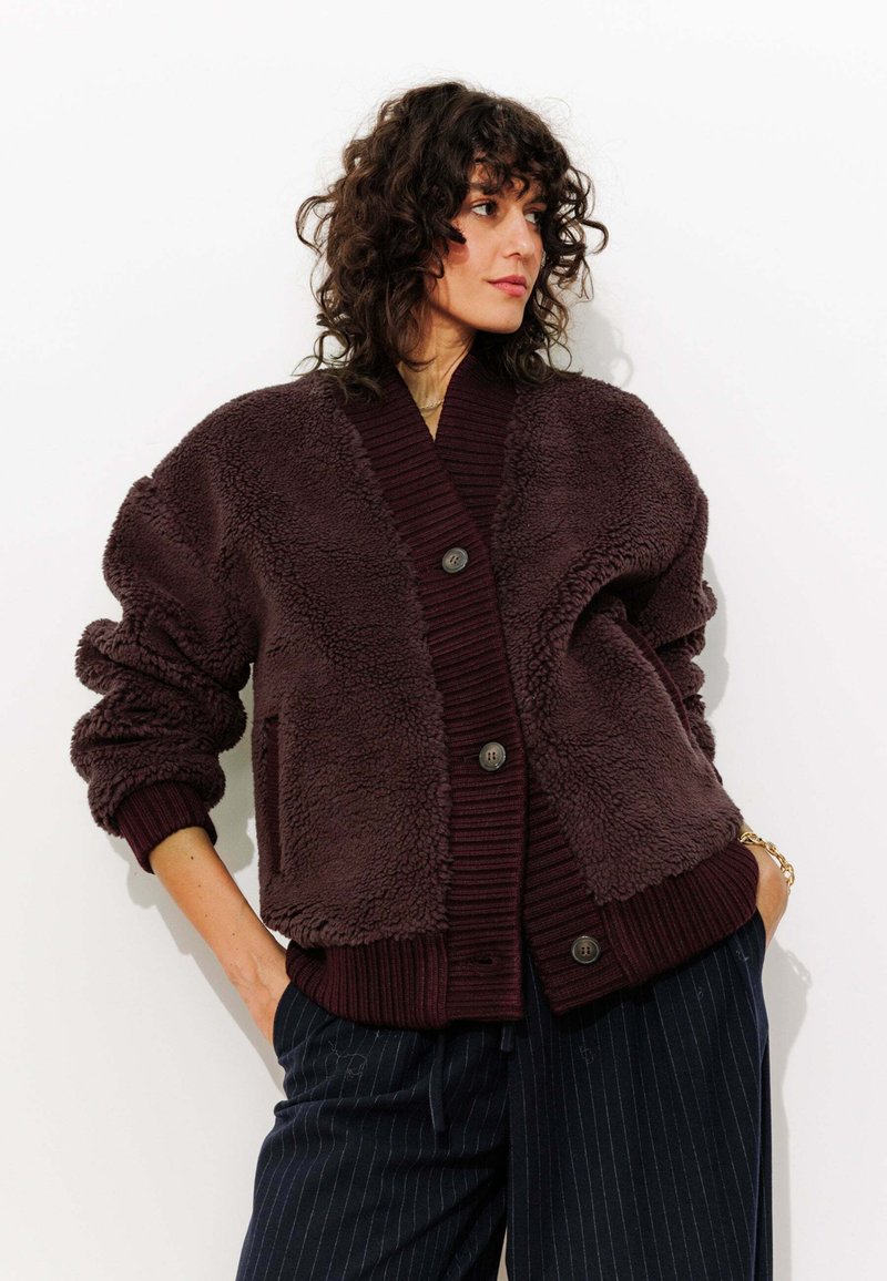 Veste en polaire marron foncé avec des poignets et un col en bordeaux côtelé. Dote de grands boutons et d'une coupe décontractée. Texture lisse et design casual.