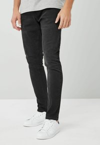 Juodi slim fit džinsai su lygiu paviršiumi, turintys klasikinį 5 kišenėlių dizainą. Suderinti su baltais sportiniais batais ir pilku marškinėliu.