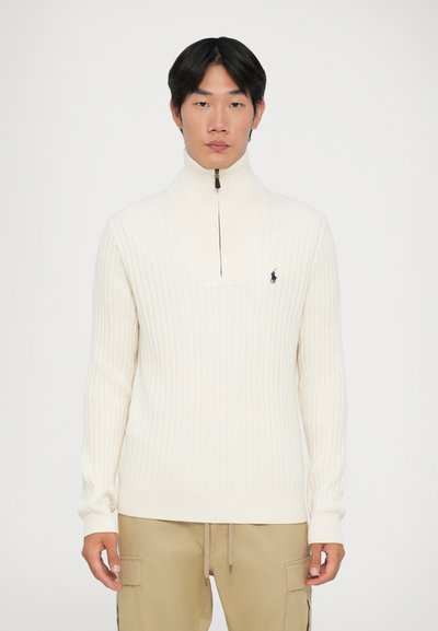 Jersey acanalado crema con cuello alto y cierre de cremallera, que presenta un pequeño logo bordado en el pecho. Llevado con pantalones beige.