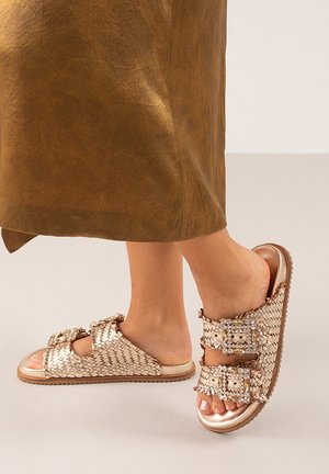 Sandalias de oro trenzadas con dos correas de hebilla y acentos decorativos. La suela es plana con una superficie de goma texturizada para mayor agarre.