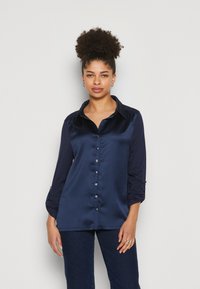 Navy Satin-Hemd mit Knopfleiste und hochgeschlagenen Ärmeln, kontrastierenden matten Ärmeln und silbernen Knopf-Details. Glatte Textur und klassisches Kragen-Design.