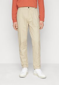 Pantaloni in cotone beige con una vestibilità comoda, pieghe frontali e chiusura a bottone. Abbinati a sneakers bianche, caratterizzate da una texture liscia e un design minimali.