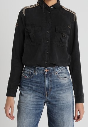 Persona que lleva una camisa negra abotonada con adornos decorativos en los hombros y dos bolsillos delanteros combinada con vaqueros azules.
