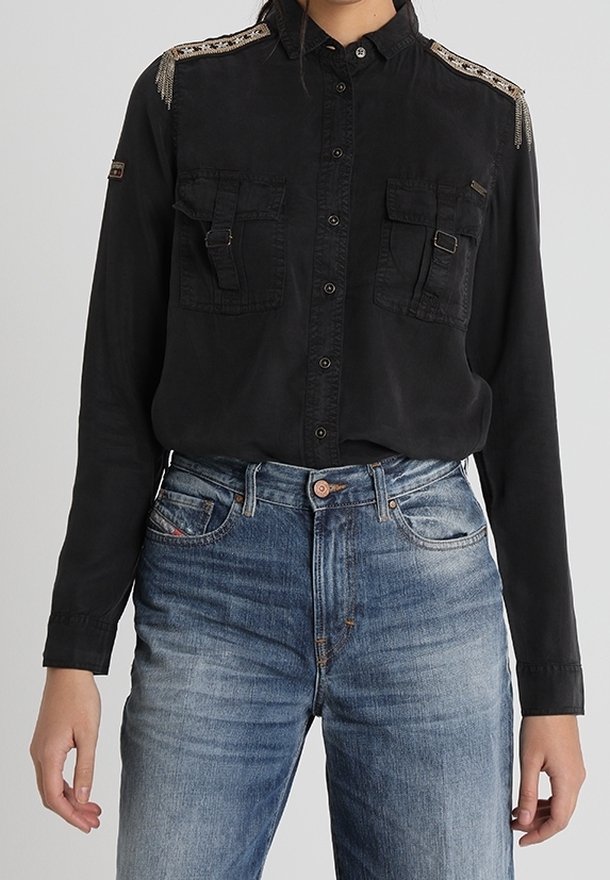 Personne portant une chemise noire à boutons avec des ornements décoratifs aux épaules et deux poches avant, associée à un jean bleu.