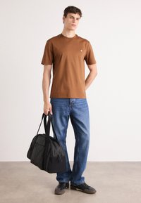 Hnedé bavlnené tričko s krátkymi rukávmi v kombinácii s modrými džínsy. Model drží čiernu látkovú tašku typu duffel s ramennými popruhmi.
