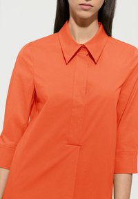 Orangenes Hemd mit Kragen und 3/4-Ärmeln, das über eine Knopfleiste und subtile Faltendetails an der Vorderseite verfügt, gefertigt aus glattem Stoff.