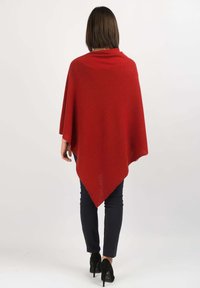 Puncho punah merah berbentuk segitiga, dengan tekstur halus dan desain terurai. Dipakai dengan celana hitam dan sepatu hak.