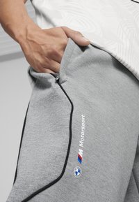 Grå sweatpants med sorte accenter, blød tekstur, med en sidelomme og "Motorsport" logoen med BMW-emblemet på låret.
