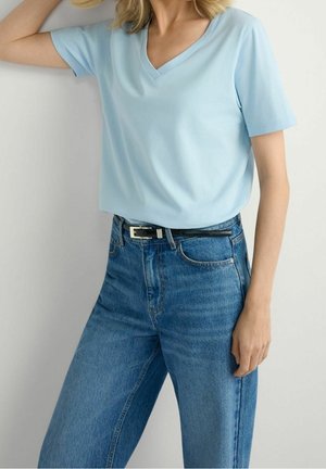 Woman wearing a light blue short-sleeve V-neck shirt tucked into high-waisted blue jeans with a black belt.