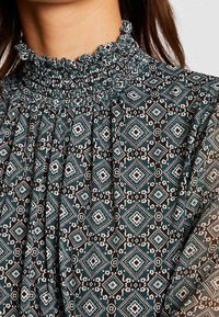 Schwarzes Stoffoberteil mit gesmoktem hohen Kragen, kompliziertem türkis-weißen geometrischen Diamantmuster mit floralen Akzenten.