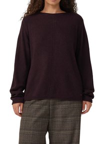 Vrouw in een donkerpaarse lange mouwen trui en grijze geruite broek, staande met ontspannen armen langs haar zij op een witte achtergrond.