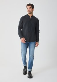Schwarz langärmliges Hemd mit Knöpfen, aus leichtem Stoff mit subtiler Textur, kombiniert mit hellblauen Jeans und schwarzen Schnürstiefeln.