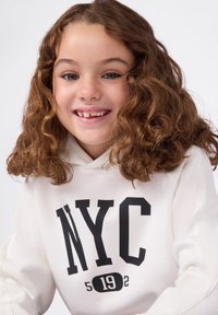 Sudadera blanca con un estampado negro de "NYC" y el año "5192". El material parece suave; el modelo tiene el cabello rizado y marrón y sonríe, mostrando un diente faltante.