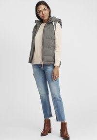 Grau gepolsterte Weste mit Kapuze, vorne mit Reißverschluss und seitlichen Taschen, über einem beigen Pullover und blauen Jeans mit braunen Stiefeletten getragen.