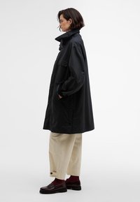 Casaco preto longo com colarinho alto e botões, exibido sobre calças largas de cor clara e mocassins escuros, apresentando bolsos laterais.