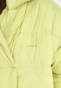 Lys grøn pufjakke med en struktureret overflade, der har et syet mønster og en diskret Calvin Klein-logo broderet på brystet.
