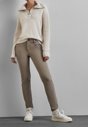 Henkilö, jolla on beigejä kapeita housuja, kermaisen värinen neulepusero vetoketjukauluksella ja valkoiset korkeavartiset tennarit, seisoo harmaan taustan edessä.