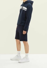 Osoba v námořnickém modrém mikině s bílými pruhy, námořnických cargo šortkách, bílých kotníkových ponožkách a bílých teniskách, stojící na světle jednobarevném pozadí.