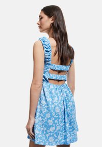 Blauwe bloemenjurk van textuurstof, met ruffles op de schouderbanden, een open rug met uitsnijdetails, en een uitlopende rok. Witte bloemenprint.