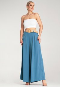 Szerokie niebieskie spodnie z zakładkami, połączone z białą górą bez ramiączek. Outfit uzupełnia beżowy pasek z okrągłą klamrą i gładką teksturą.
