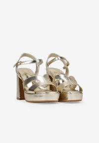 Metallisch goldene Plateausandalen mit einem klobigen Absatz, ausgestattet mit mehreren Riemen und einem Schnallenverschluss. Glatte Oberfläche, minimale Details.