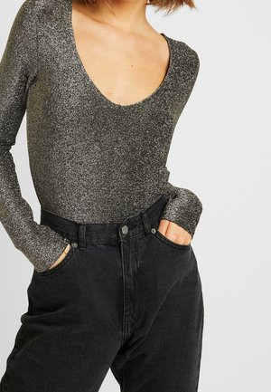 Femme portant un haut scintillant à manches longues avec un col rond et un jean noir ample taille haute, les mains dans les poches.