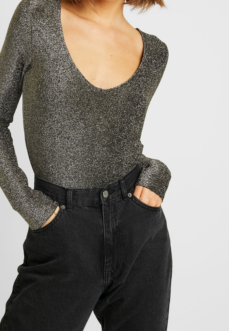 Femme portant un haut scintillant à manches longues avec un col rond et un jean noir ample taille haute, les mains dans les poches.