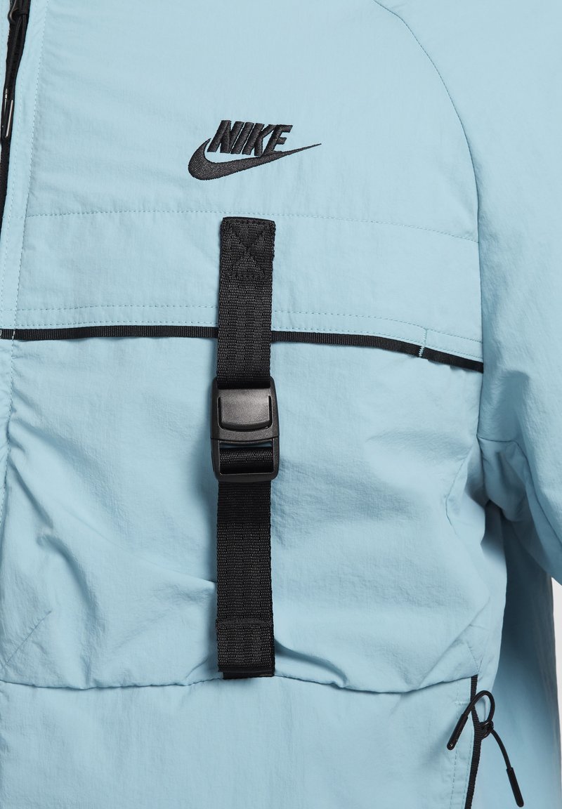 Jasnoniebieska kurtka przeciwdeszczowa z czarnym logo Nike, regulowany czarny pasek z klamrą oraz kontrastowe czarne wykończenia wzdłuż klatki piersiowej.