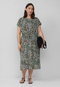 Musterkleid mit kurzen Ärmeln und tailliertem Gürtel, das ein geometrisches Design in Schwarz, Weiß, Türkis und Braun aufweist, kombiniert mit einer schwarzen Tasche.