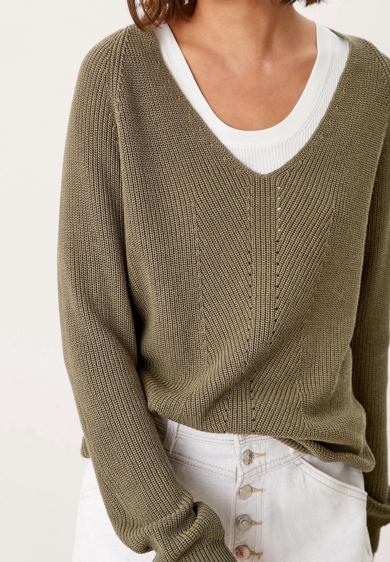 Femme portant un pull en maille vert olive texturé à col en V par-dessus un top blanc côtelé, associé à un pantalon blanc à taille haute avec fermeture à boutons.