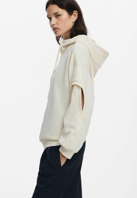 Cream hoodie with a relaxed fit, featuring a large hood, side openings in the sleeves, and ribbed cuffs. Worn with dark pants.
