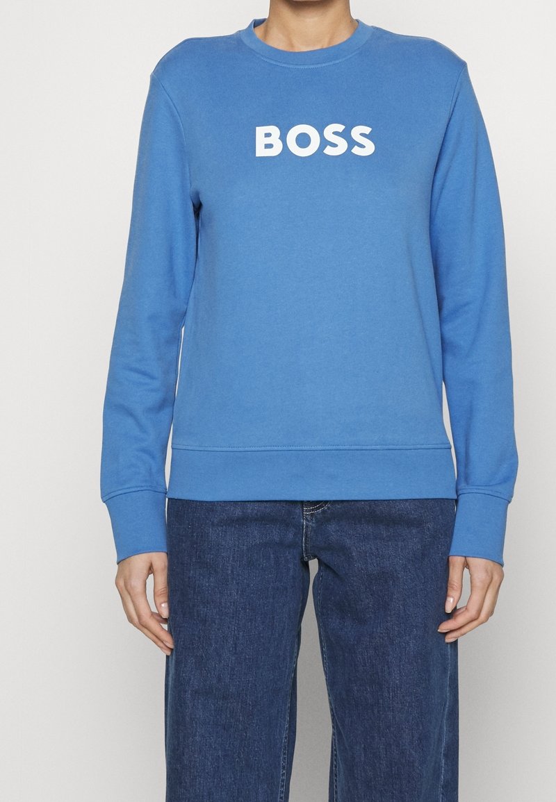 Person som bär en blå sweatshirt med "BOSS" i vit text på bröstet och mörkblå jeans mot en neutral bakgrund.