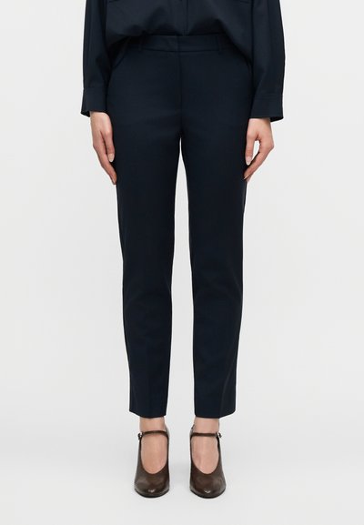 Femme portant un pantalon slim bleu marine et des talons Mary Jane marron texturés, debout devant un fond clair uni.