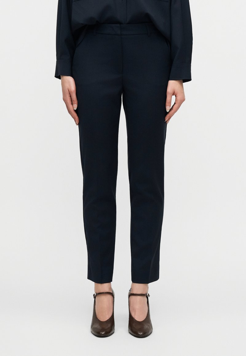 Γυναίκα που φοράει στενά σκούρα μπ λε παντελόνια και καφέ υφασμάτινες γόβες Mary Jane, που στέκεται μπροστά σε ένα απλό φωτεινό φόντο.