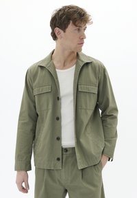Olivengrüne, leichte Jacke mit Kragen, zwei Brusttaschen, Knopfverschluss und langen Ärmeln. Kombiniert mit einem weißen, gerippten Tanktop.