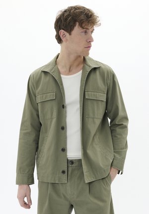 Olive green, lightweight jacket with a collar, two chest pockets, button closure, and long sleeves. Paired with a white ribbed tank top.