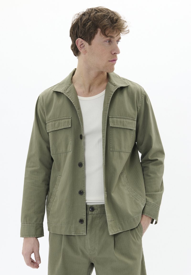 Olivengrüne, leichte Jacke mit Kragen, zwei Brusttaschen, Knopfverschluss und langen Ärmeln. Kombiniert mit einem weißen, gerippten Tanktop.