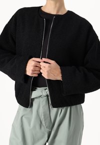 Giacca bomber in pile nero con scollatura rotonda, chiusura con zip e vestibilità ampia. Presenta maniche larghe e un tessuto testurizzato. Abbinata a pantaloni verde menta.