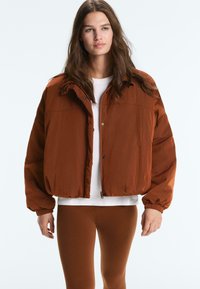 Ženska z dolgimi rjavimi lasmi, oblečena v rjavo rjavo puhasto jakno, belo majico in ujemajoče se rjave legice, stoji pred enobarvnim ozadjem.