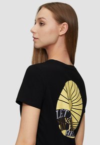 Zwarte katoenen T-shirt met een grote, gele spiraalafbeelding op de achterkant en tekst die luidt: "LAAT DE ZON IN JE HART SCHIJNEN."