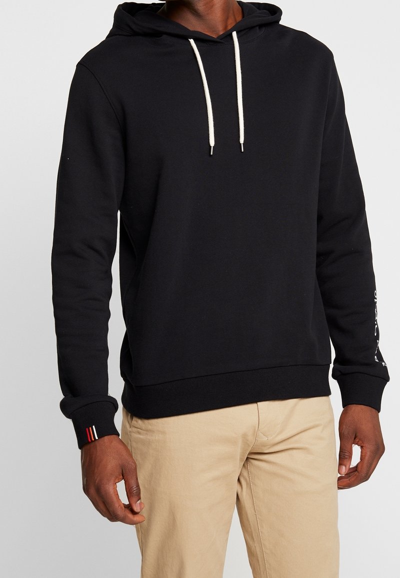 Svart hoodie med vita dragsnören, ribbade ärmslut och ett solid nederkant; har små accentstripes i röd, vit och blå på en ärm.