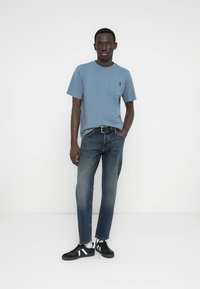 Ανοιχτό μπλε κοντομάνικο μπλουζάκι με τσέπη, συνδυασμένο με σκούρα μπλε τζιν και μαύρα αθλητικά παπούτσια με λευκές ρίγες.